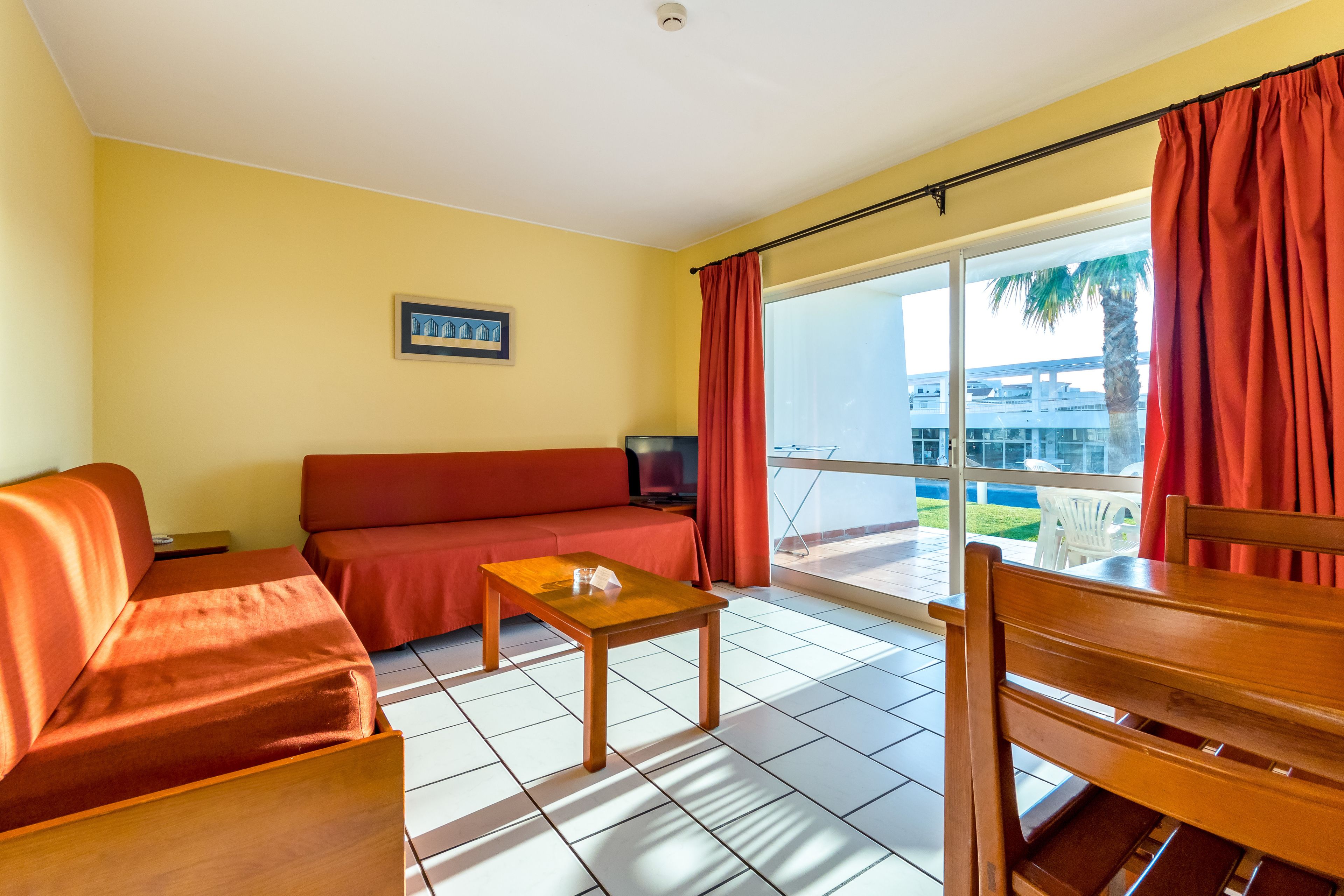 A room with a sofa, a small table, and a chair. There is a window with curtains and a view of palm trees and buildings outside.
