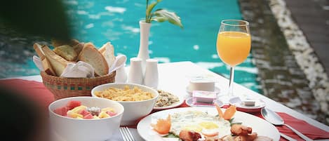 A breakfast setting by a pool with a plate of eggs, bacon, and toast, a bowl of fruit, and a glass of orange juice.