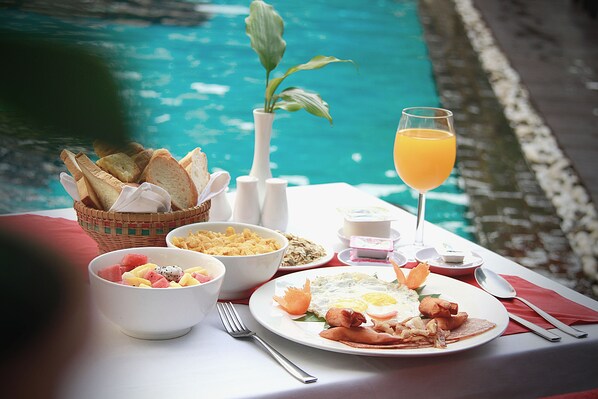 A breakfast setting by a pool with a plate of eggs, bacon, and toast, a bowl of fruit, and a glass of orange juice.