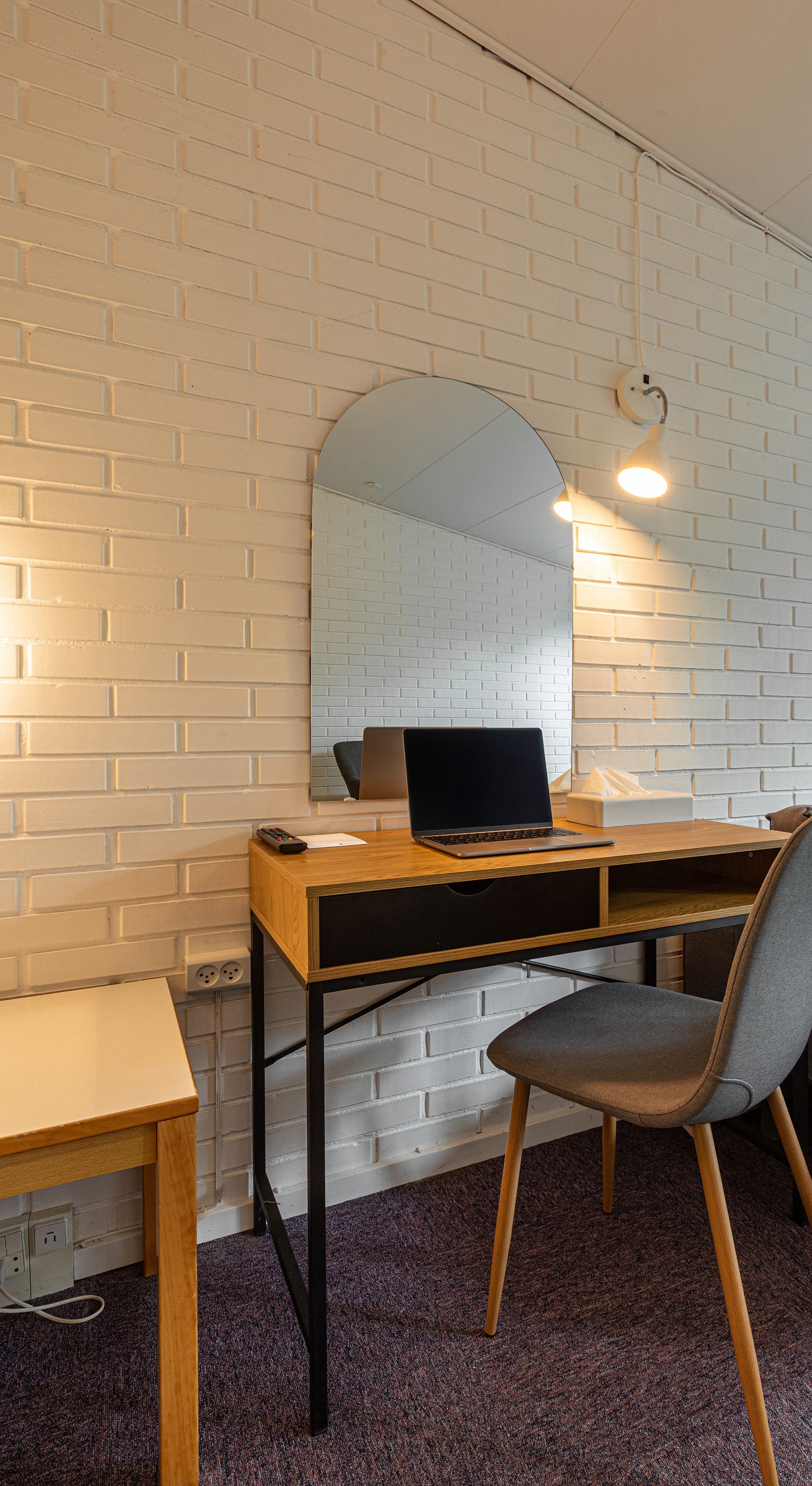 A room with a white brick wall, a wooden desk with a laptop, a chair, and a mirror.