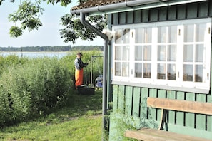 Ein rustikales Holzgebäude mit einer Bank, umgeben von üppigem Grün und einem Wasserspiegel im Hintergrund.