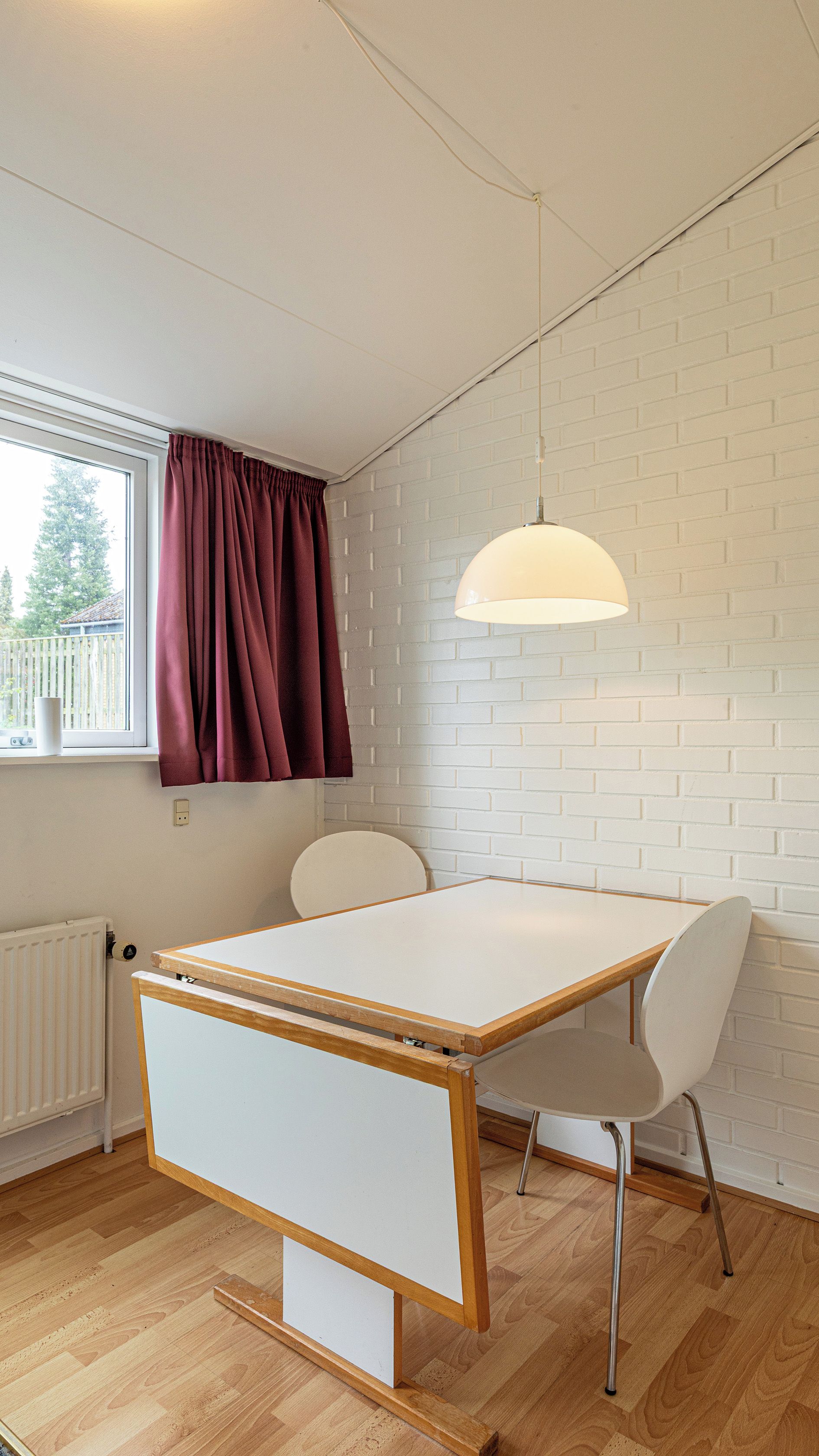 A room with a white table, two chairs, a window with curtains, a radiator, and a hanging light fixture.