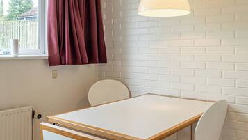 A room with a white table, two chairs, a window with curtains, a radiator, and a hanging light fixture.