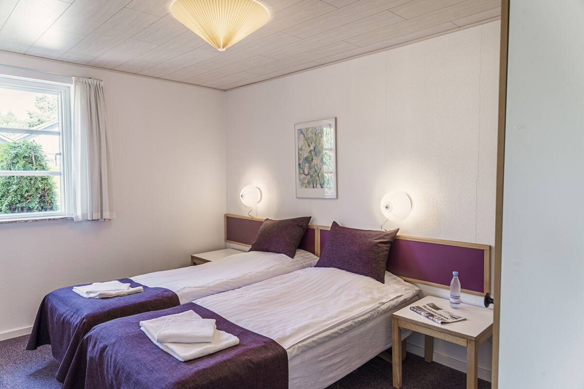 Une chambre d’hôtel avec deux lits, une fenêtre avec des rideaux, une œuvre d’art accrochée au mur, des lampes de chevet, une table de chevet avec une bouteille d’eau et un magazine, et un luminaire au plafond.