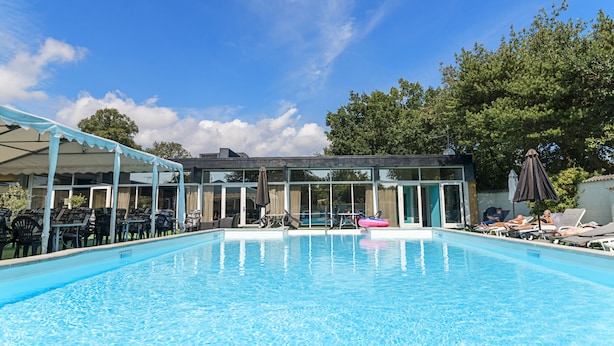 Piscina com espreguiçadeiras e guarda-sóis, cercada por uma área de estar coberta e um edifício com amplas janelas.