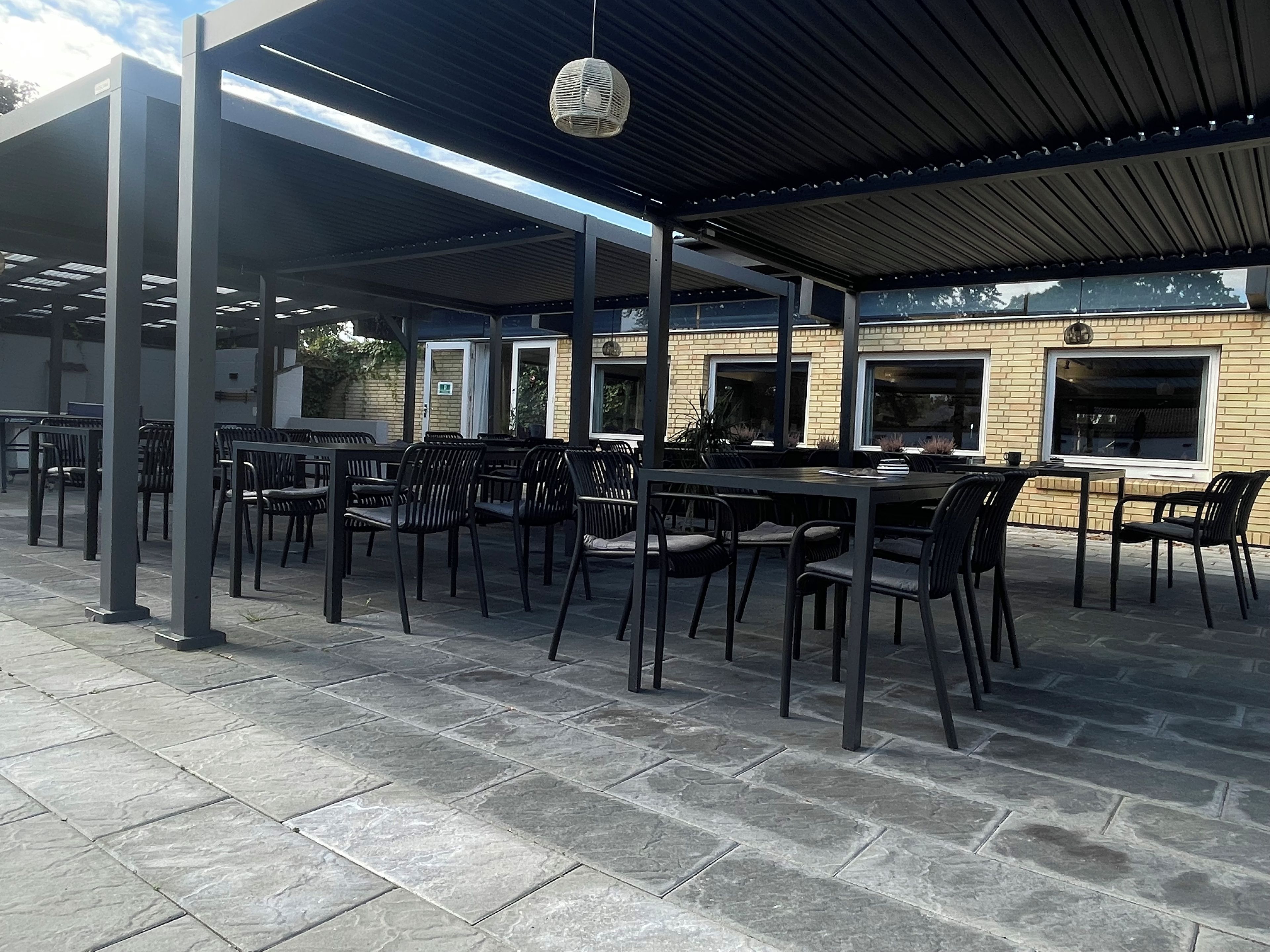An outdoor patio area with tables and chairs, a covered seating area, and a brick building in the background.