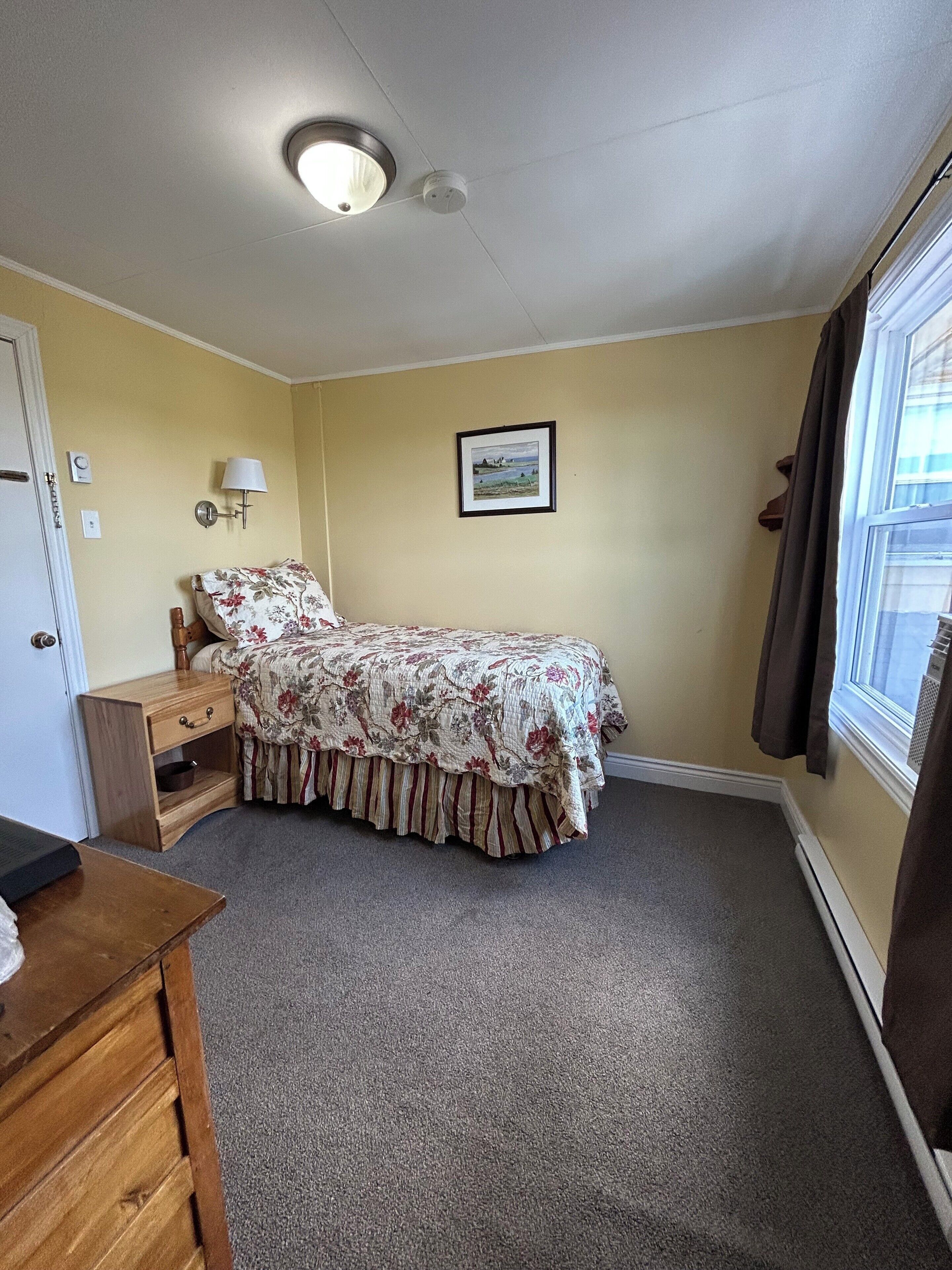A bedroom with a bed, a nightstand, a lamp, a framed picture on the wall, and a window with curtains.