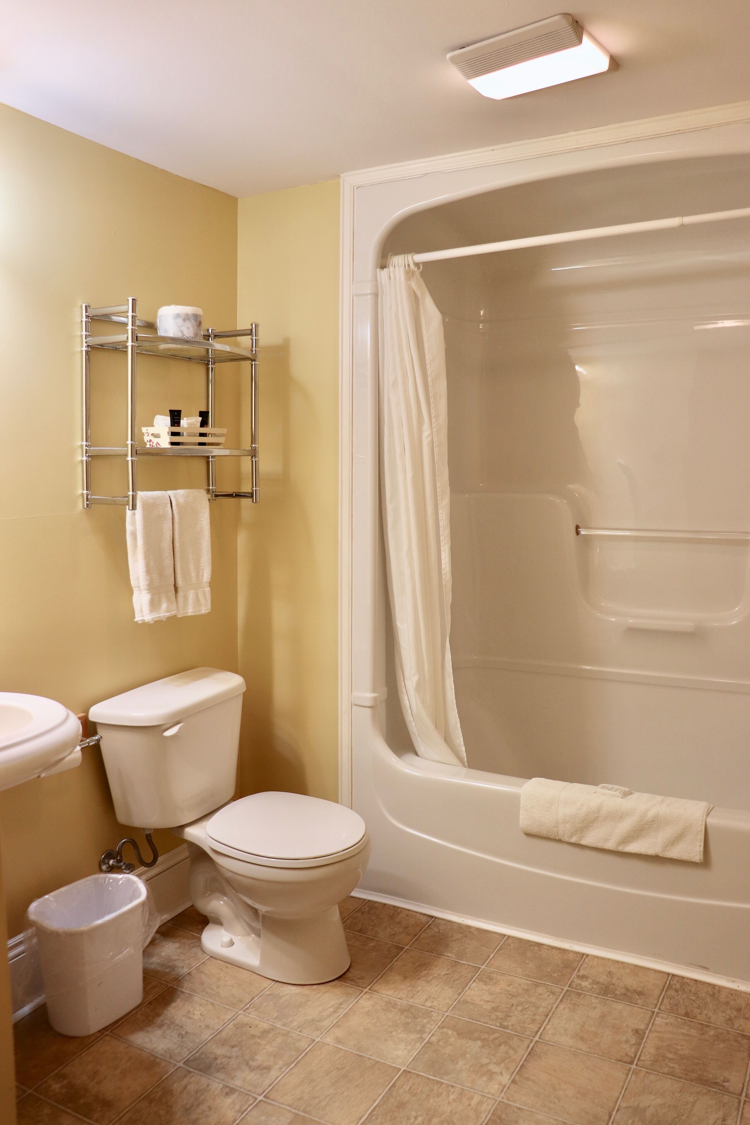 A bathroom with a toilet, sink, and bathtub. There is a shelf with toiletries and a trash can.