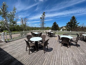 Une terrasse en bois avec des espaces de détente en plein air, comprenant des tables et des chaises rondes, le tout entouré d’une balustrade blanche.