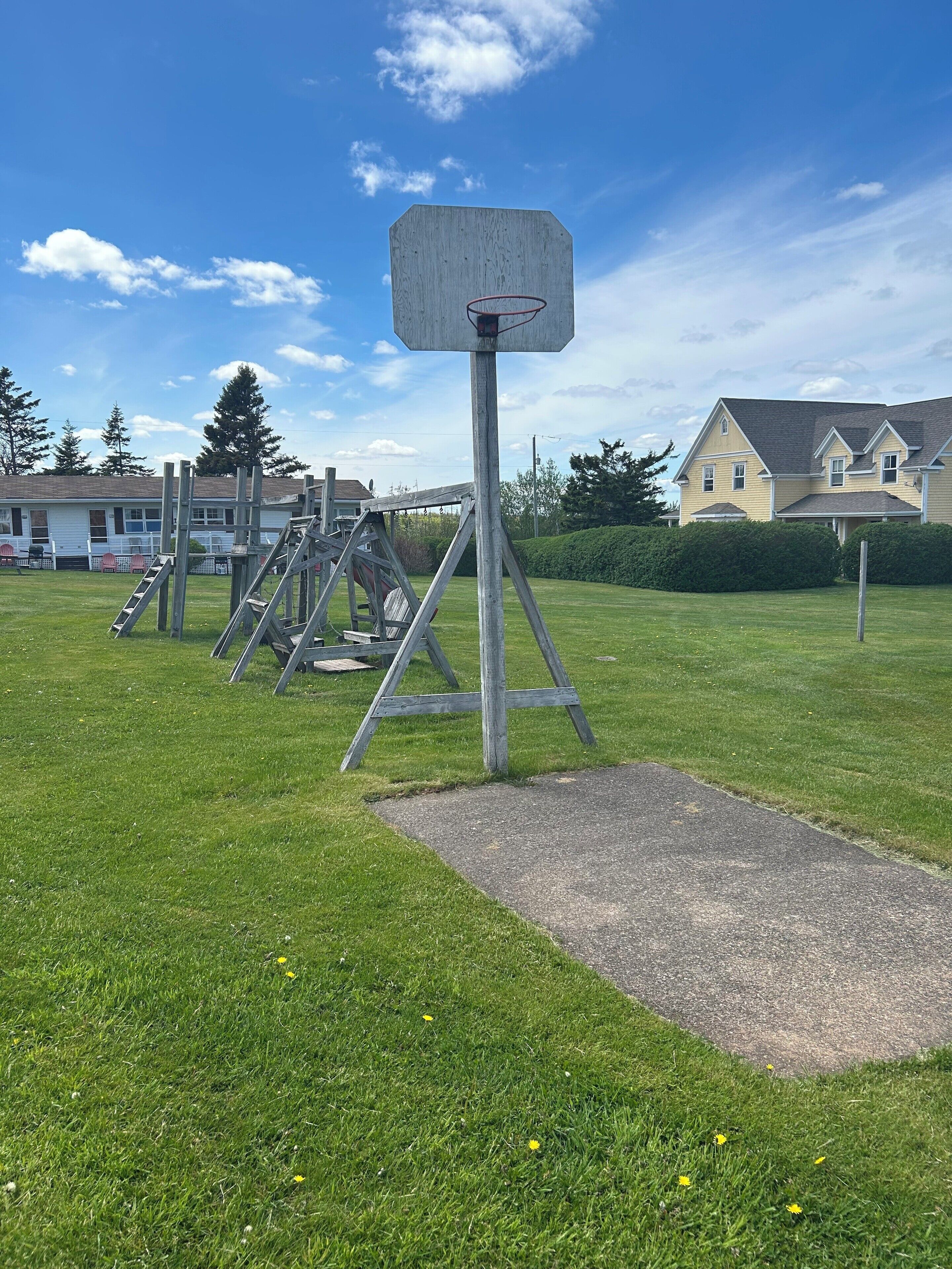 Un terrain de basket avec un panier en bois et un panneau en métal. On aperçoit en arrière-plan des gradins en métal et une zone herbeuse avec un chemin pavé.