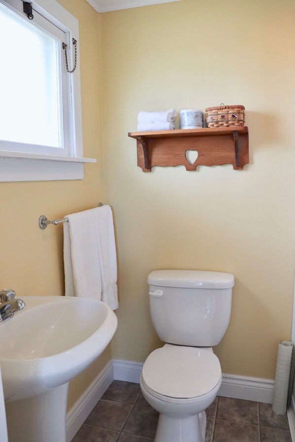 A bathroom with a white sink, toilet, and a wooden shelf above the toilet holding toiletries.