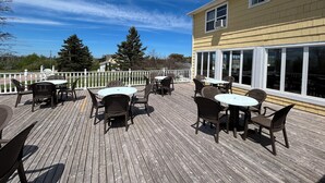 An outdoor deck with multiple tables and chairs, a white fence, and a yellow building with large windows. - Shining Waters Country Inn (Cavendish)