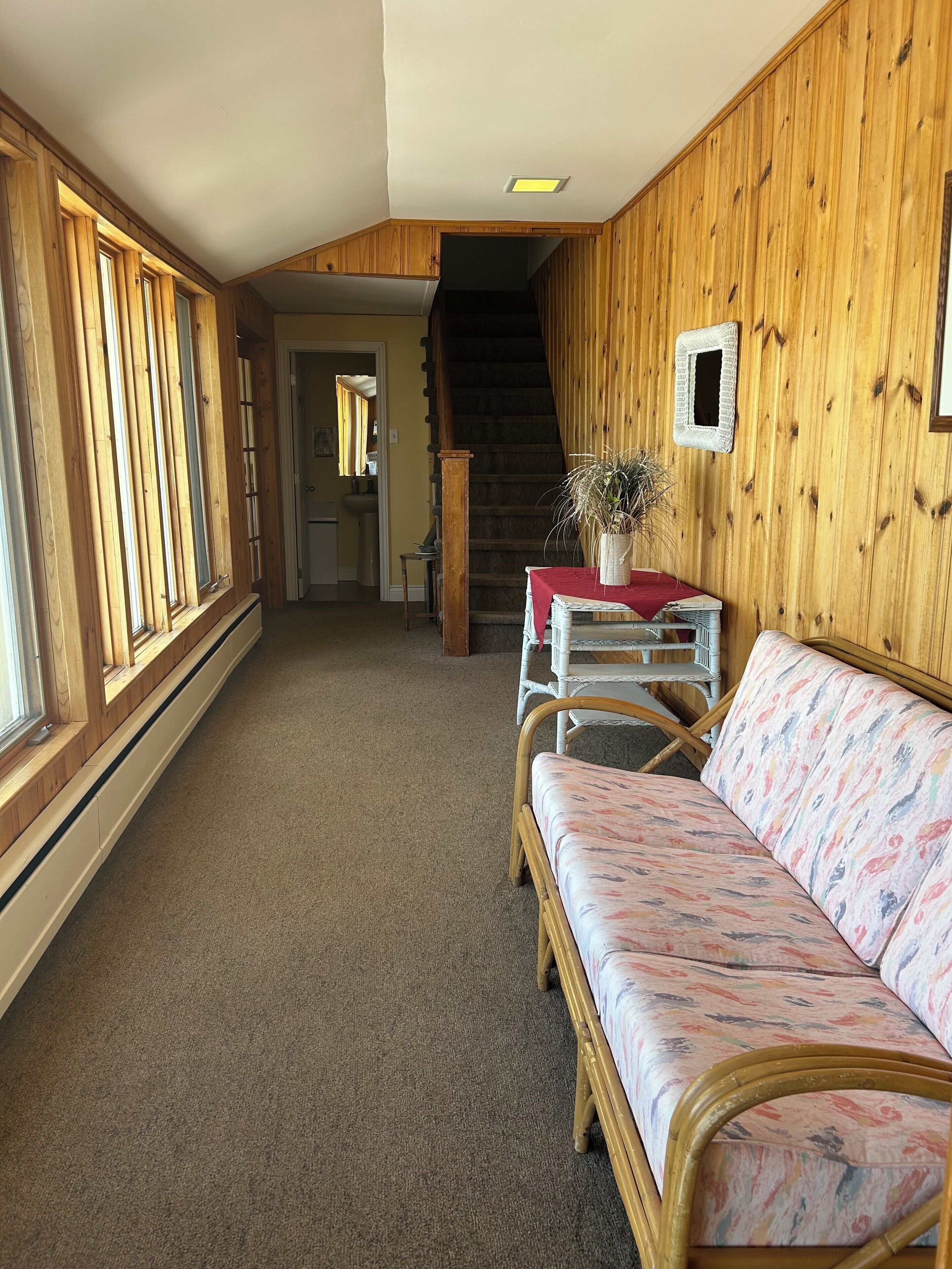 A cozy interior with a wooden wall, a sofa, a table with a red tablecloth, and a staircase leading to another room.