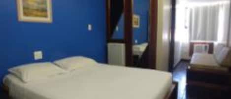 Ein Hotelzimmer mit einem Bett, einem kleinen Tisch und einem Stuhl. Ein Fenster mit Vorhängen und eine Tür zu einem anderen Raum.