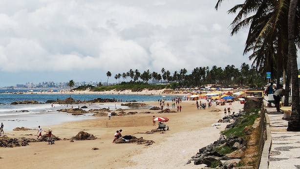 Ein Strand mit Menschen, Sonnenschirmen und einer Stadtkulisse im Hintergrund.