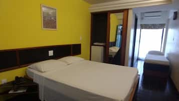 Een eenpersoonsbed in een kamer met gele wanden, een houten hoofdbord, een nachtkastje met een telefoon en een klein raam met uitzicht.