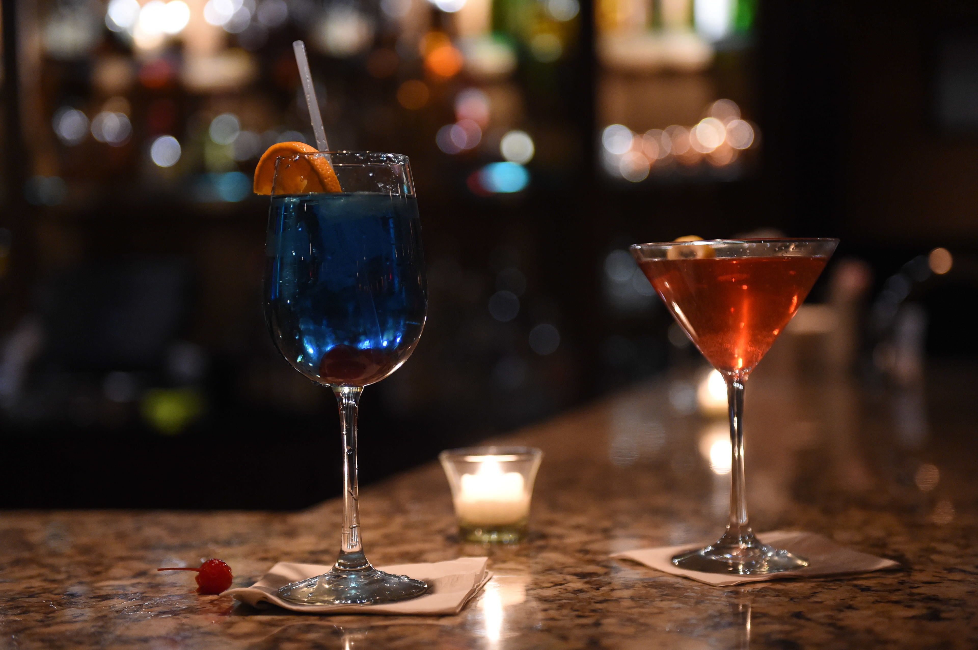 Two cocktails on a bar counter with a blurred background of bottles.