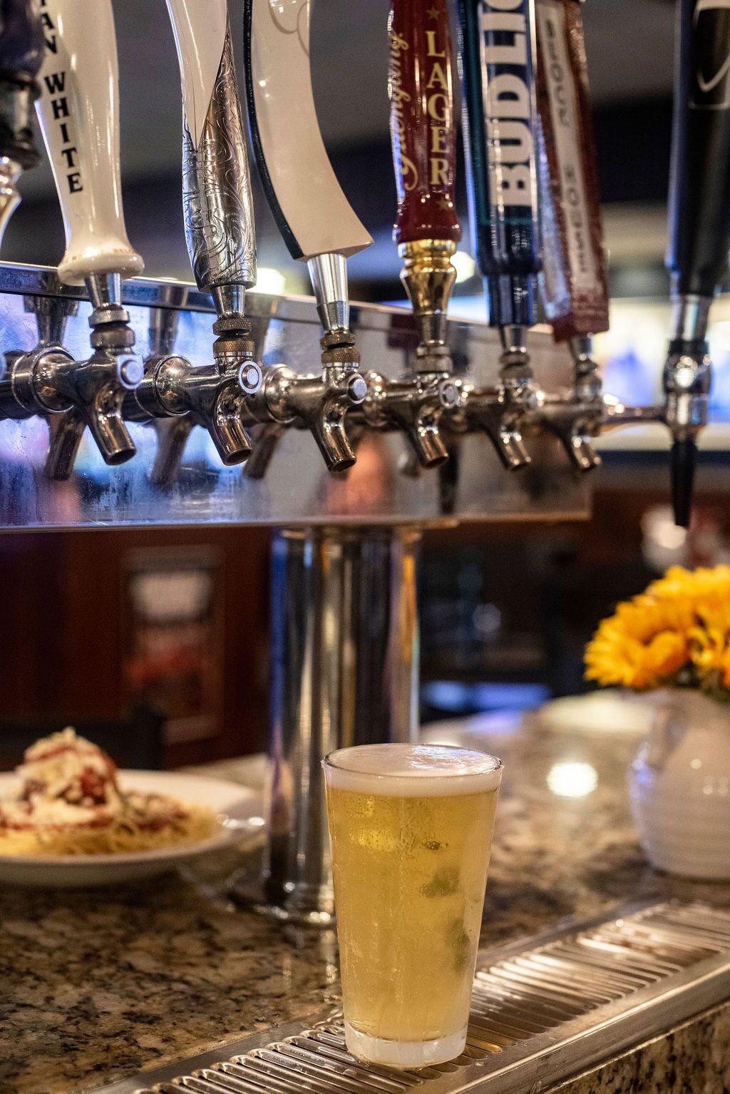 A bar area with multiple beer taps labeled with different beer types, a glass of beer, and a plate of food in the background.