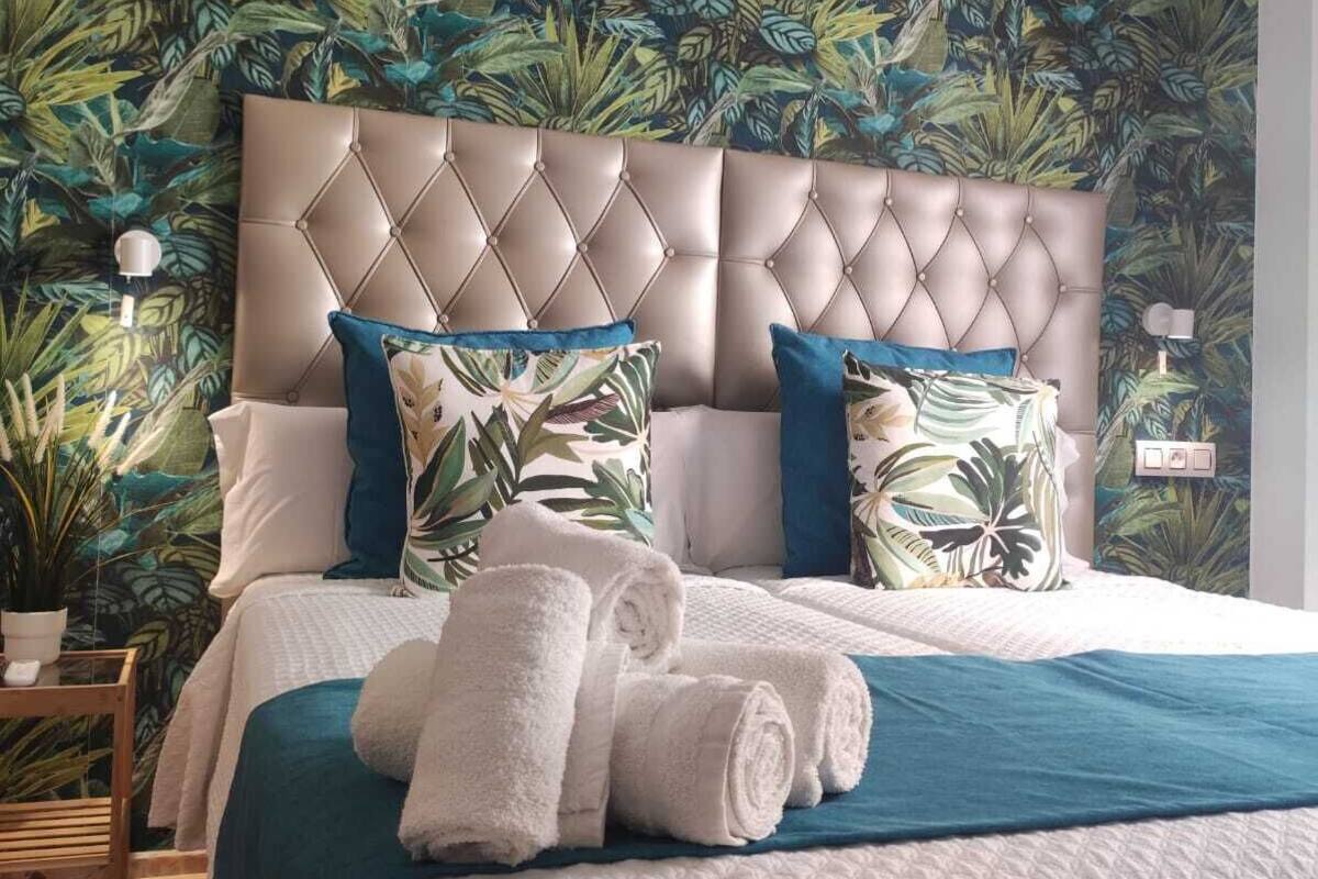 A bedroom with a large bed, a tufted headboard, and a tropical leaf-patterned wallpaper.