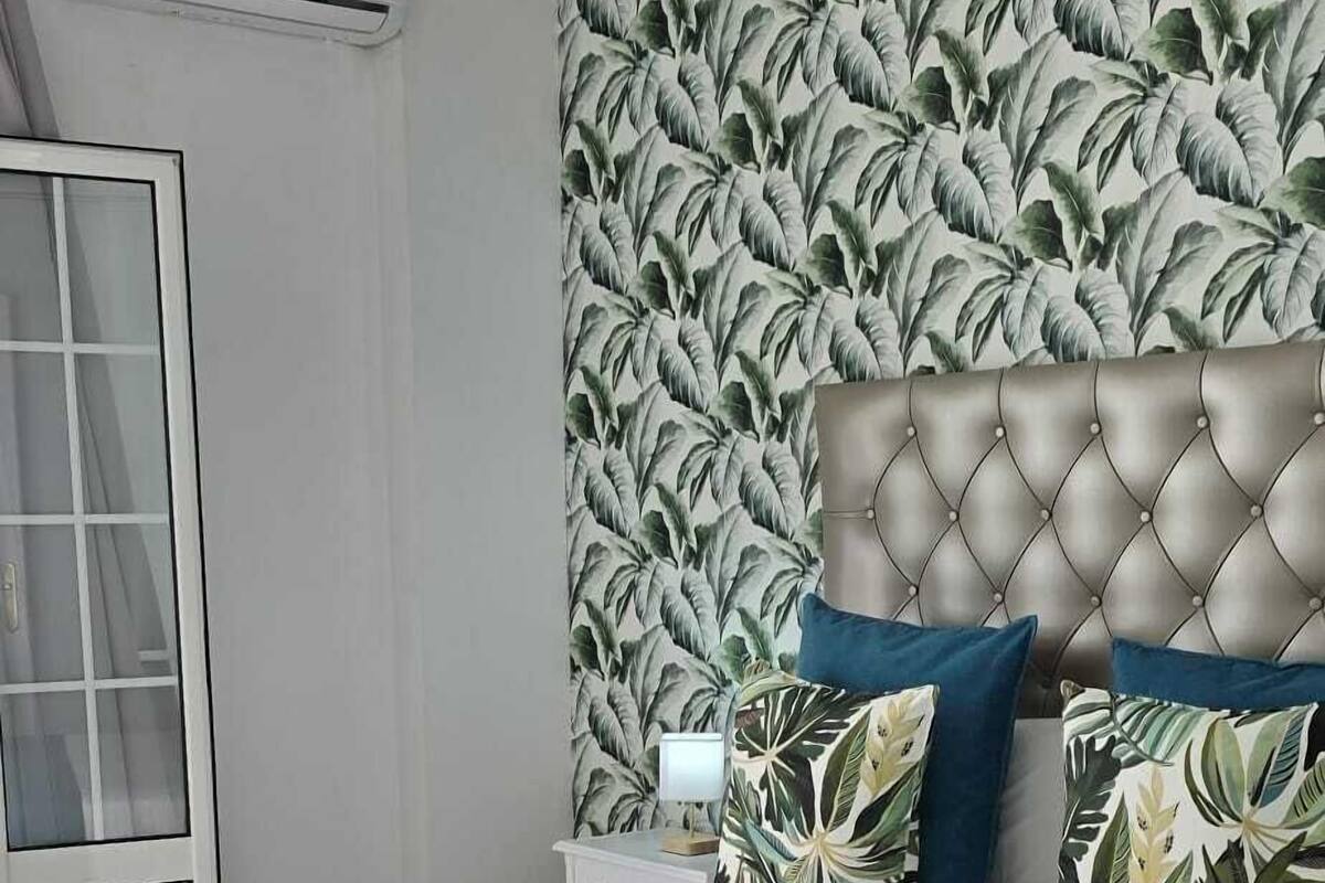 A bedroom with a tufted headboard, a bed with a blue and white bedspread, and a tropical leaf-patterned wallpaper.