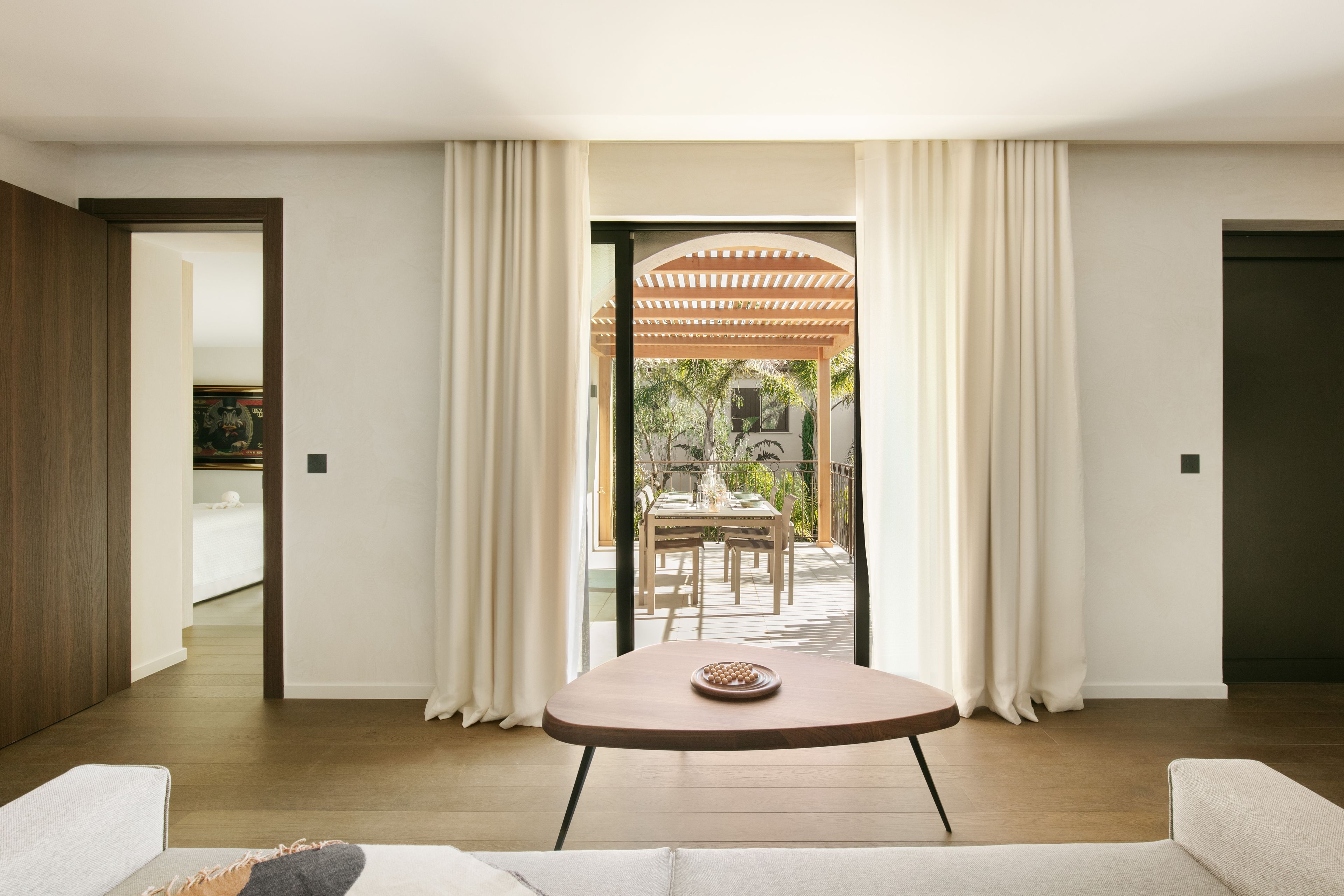 A modern living room with a round table, a sofa, and a view of an outdoor patio with a dining table and chairs.