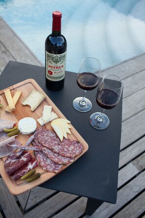 Una mesa de madera con una tabla de embutidos, dos copas de vino tinto y una botella de Petrus.