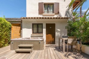 Una terraza de madera con un jacuzzi, una mesa y sillas, rodeada de follaje y un edificio con puerta y ventana.