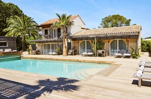 Zona de piscina con sillones reclinables, una terraza de madera y un edificio con techo de tejas.