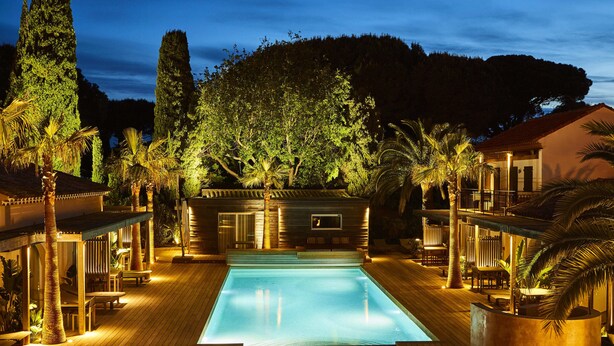 Zona de piscina con cubierta de madera, sillones de descanso y un edificio con balcón con vistas a la piscina.