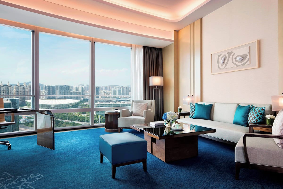 Habitación de hotel moderna con un ventanal amplio que ofrece vistas a la ciudad, un sofá, una mesa de centro y un escritorio con una silla.