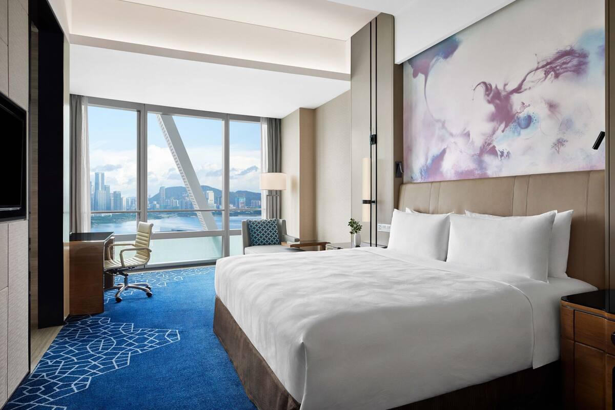 Habitación de hotel con una cama grande, un escritorio, una silla, vista de la ciudad a través de amplias ventanas y un cuadro en la pared.