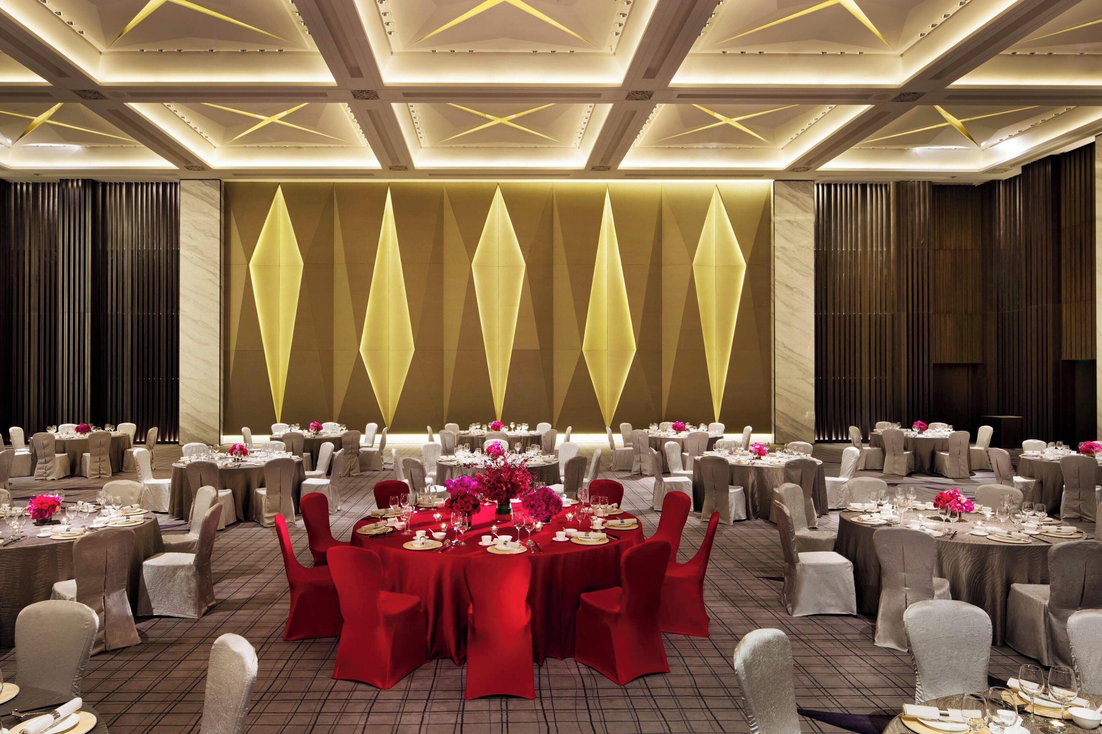 Une salle de banquet avec des tables rondes dressées pour un événement formel, comprenant une table centrale avec des chaises rouges et des arrangements floraux.