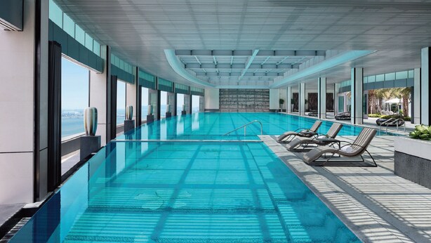 Una piscina interior moderna con agua azul cristalina, grandes ventanales y tumbonas a un lado.