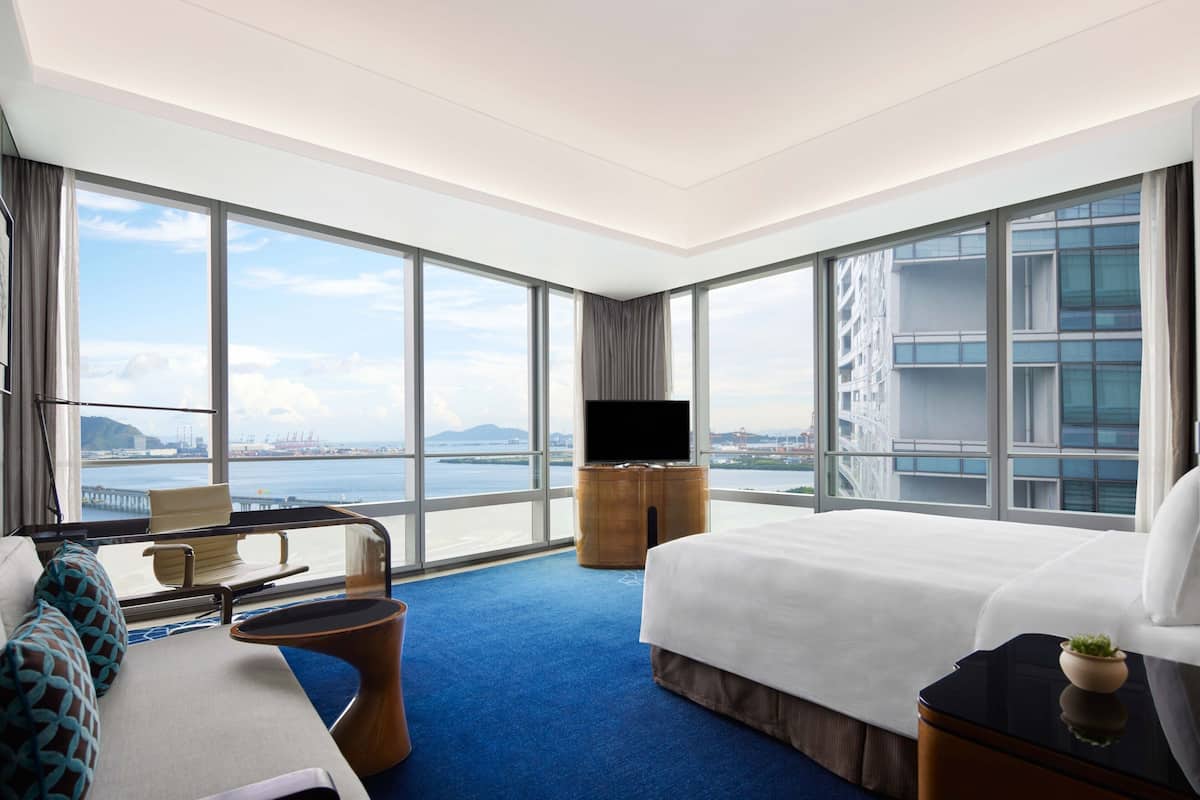 Una habitación de hotel moderna con una cama grande, televisión, zona de estar con vistas a la ciudad y balcón con vistas al agua y edificios.