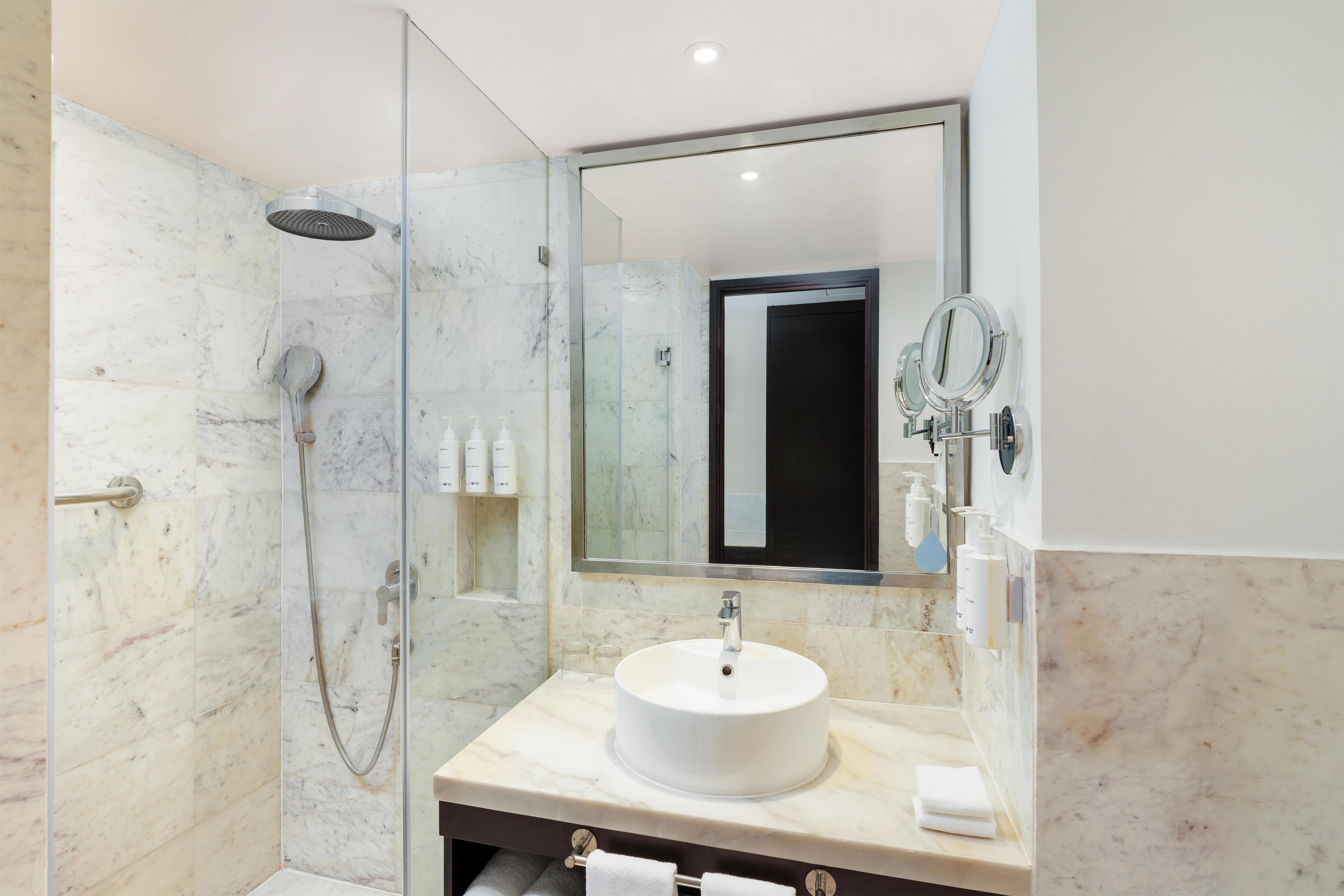 A modern bathroom with a glass-enclosed shower, a marble-tiled wall, a white sink, and a large mirror.