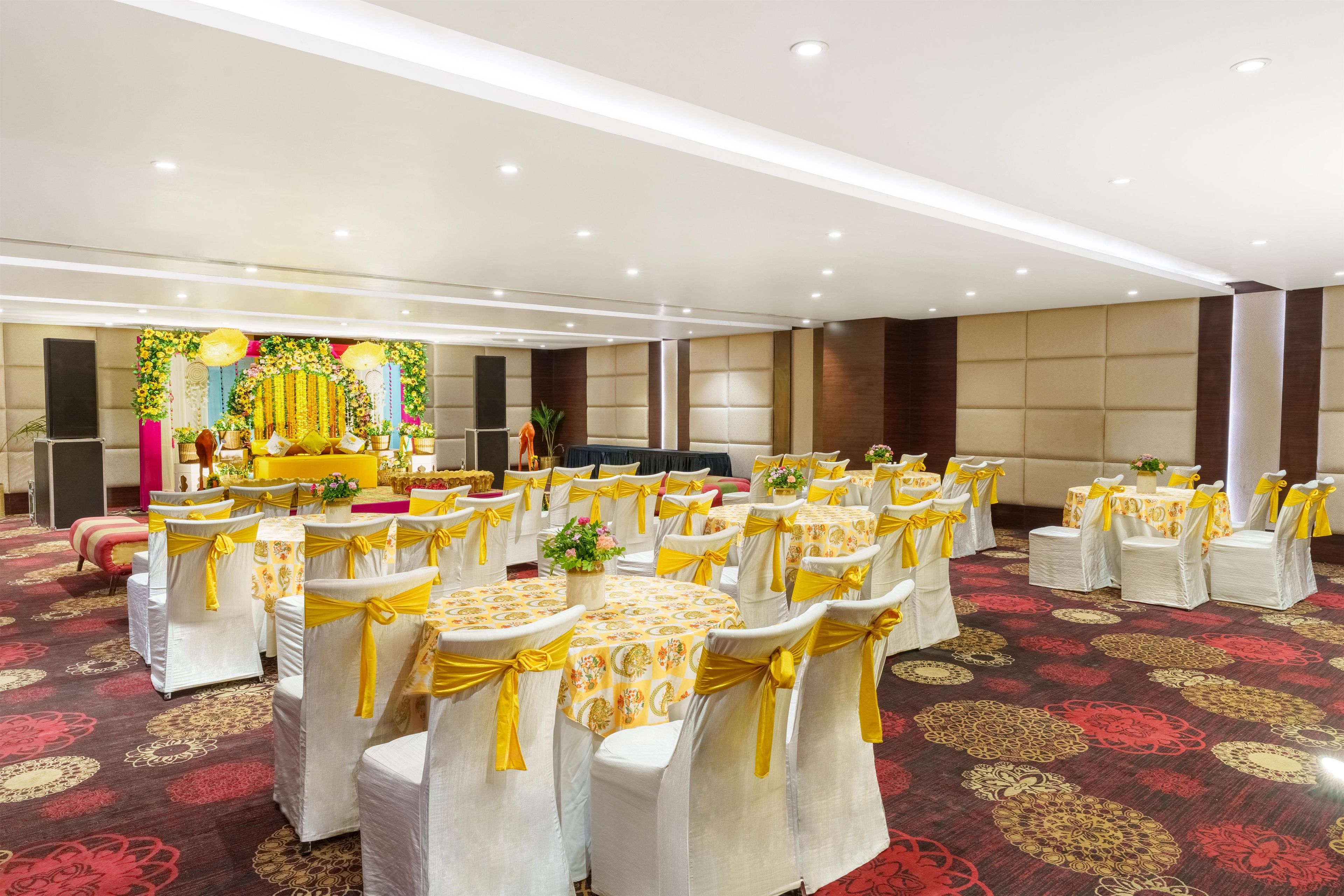 Une salle de banquet avec des tables rondes, des chaises ornées de rubans jaunes et une scène décorée de fleurs.