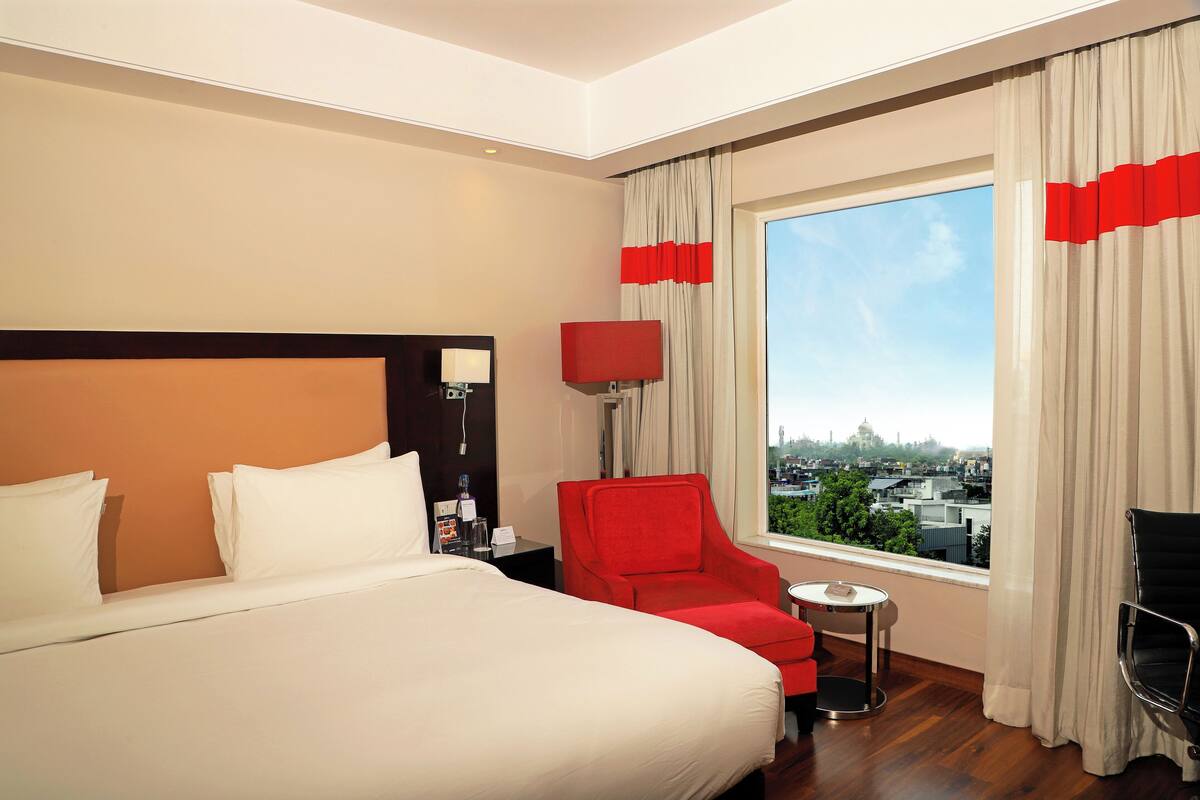 Habitación de hotel con una cama grande, un sillón rojo, una mesita y vistas a la ciudad a través de un ventanal amplio.