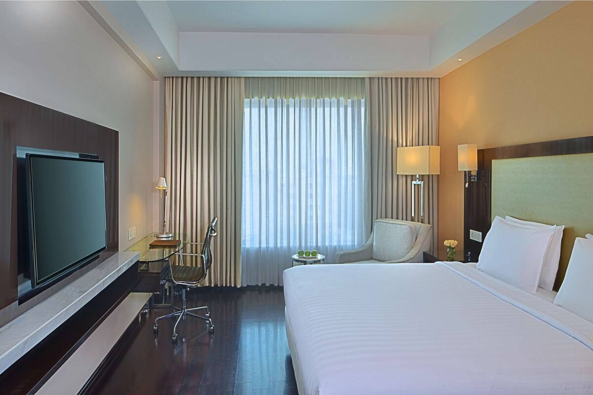 Habitación de hotel con una cama grande, televisor, escritorio con silla, mesita con lámpara y ventana con cortinas.