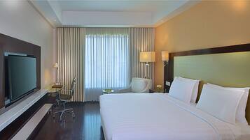 Hotelkamer met een groot bed, een televisie, een bureau met stoel, een klein tafeltje met lamp en een raam met gordijnen.