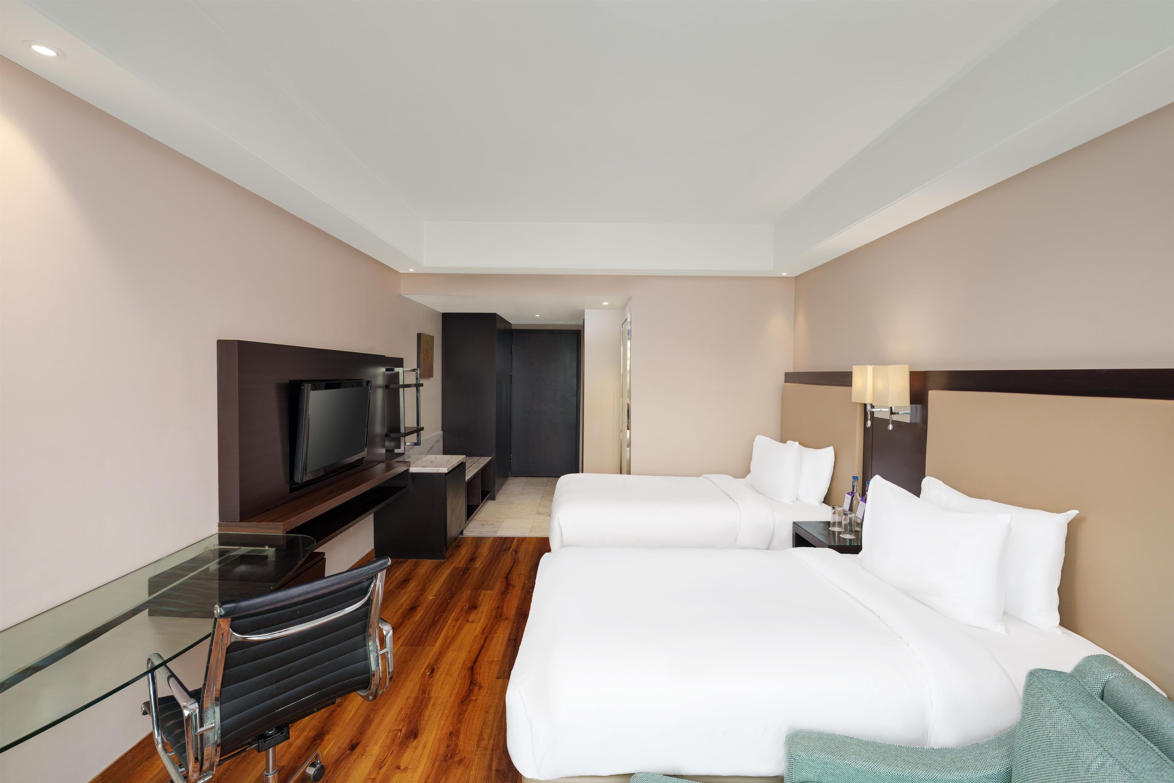 A hotel room with two beds, a TV, a desk with a chair, and a door leading to another room.