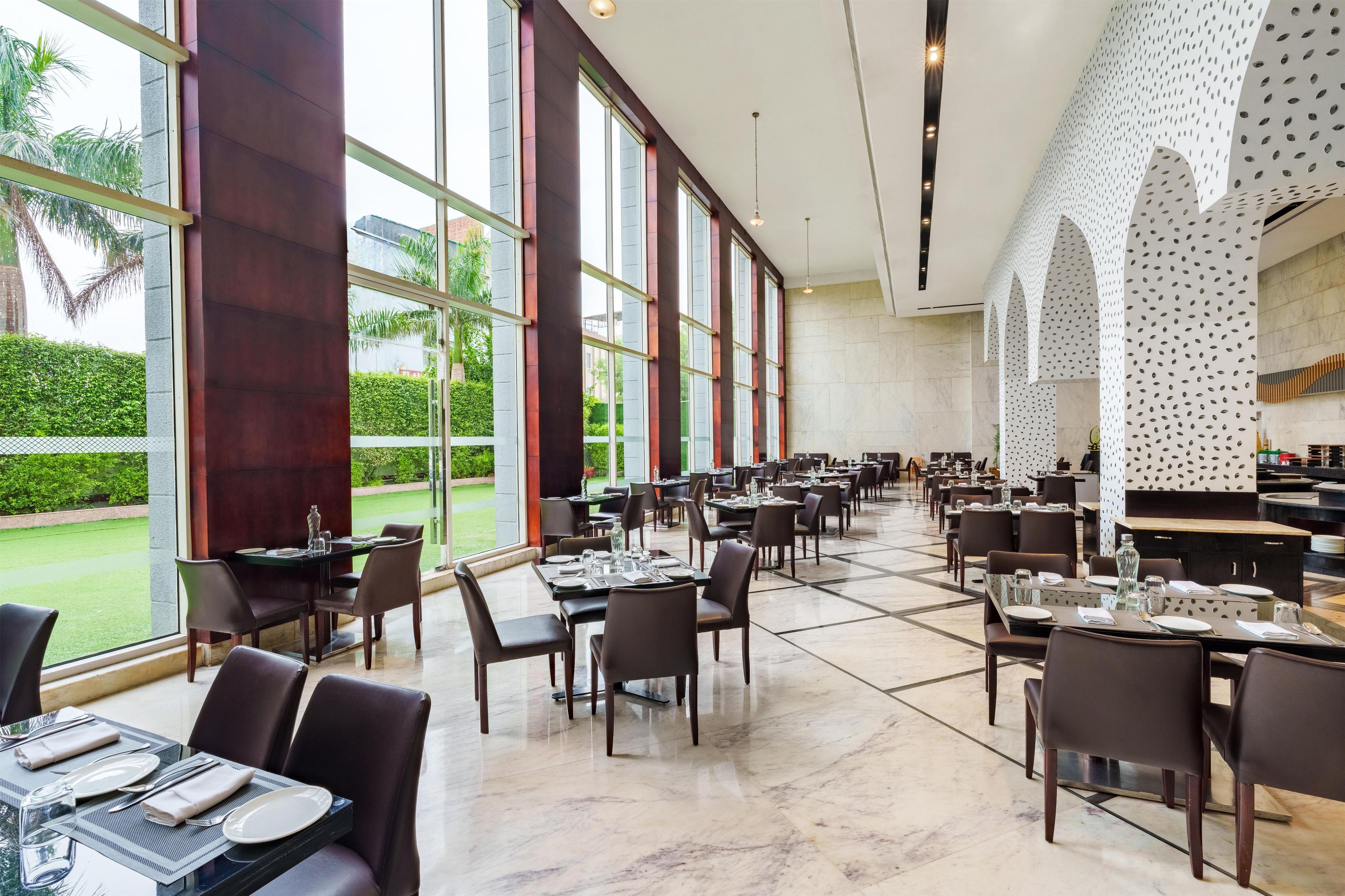 A spacious dining area with multiple tables and chairs, large windows offering a view of the outdoors, and a modern architectural design with a patterned wall.