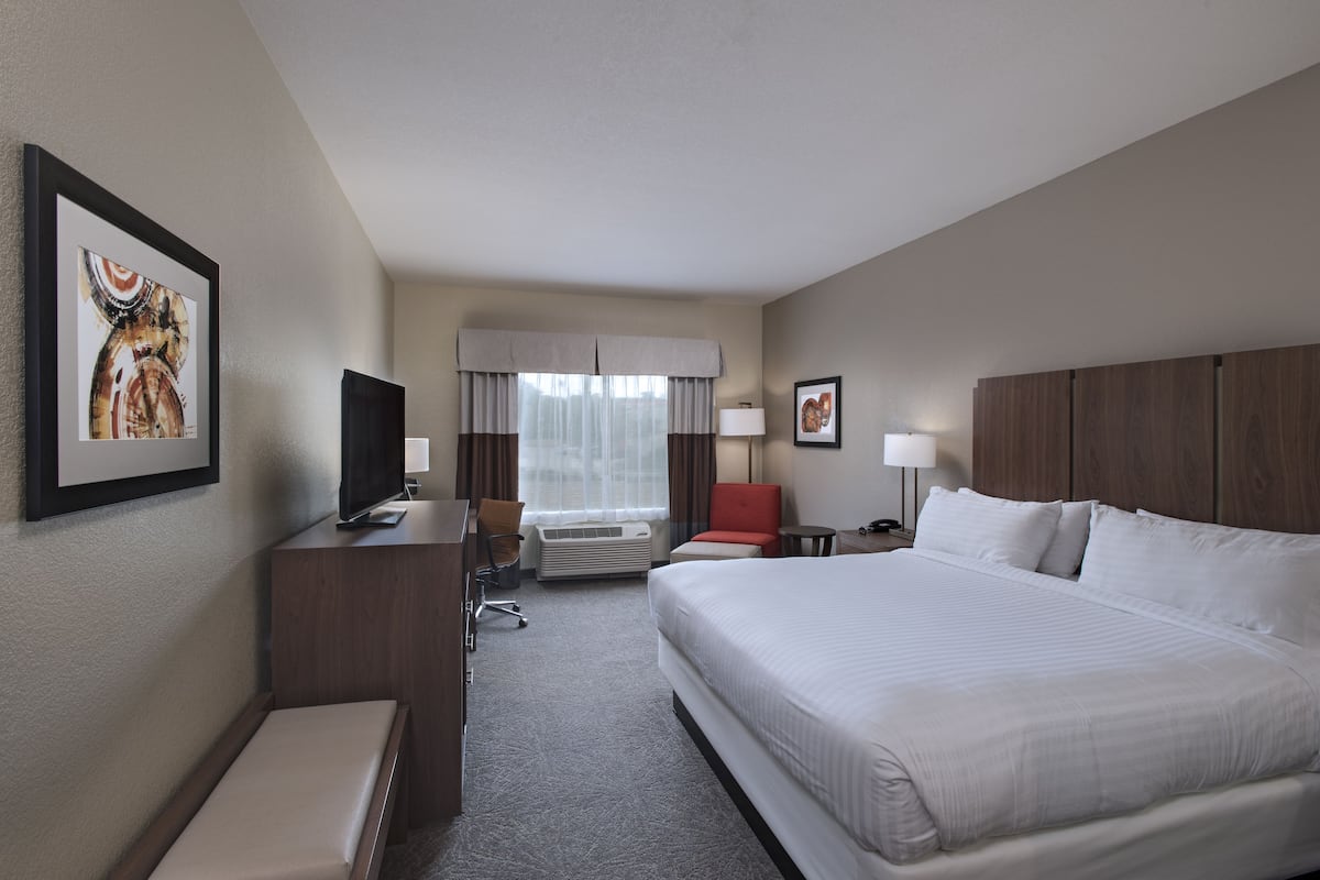 Habitación de hotel con una cama grande, un escritorio con televisor, una silla, una mesita y una ventana con cortinas.