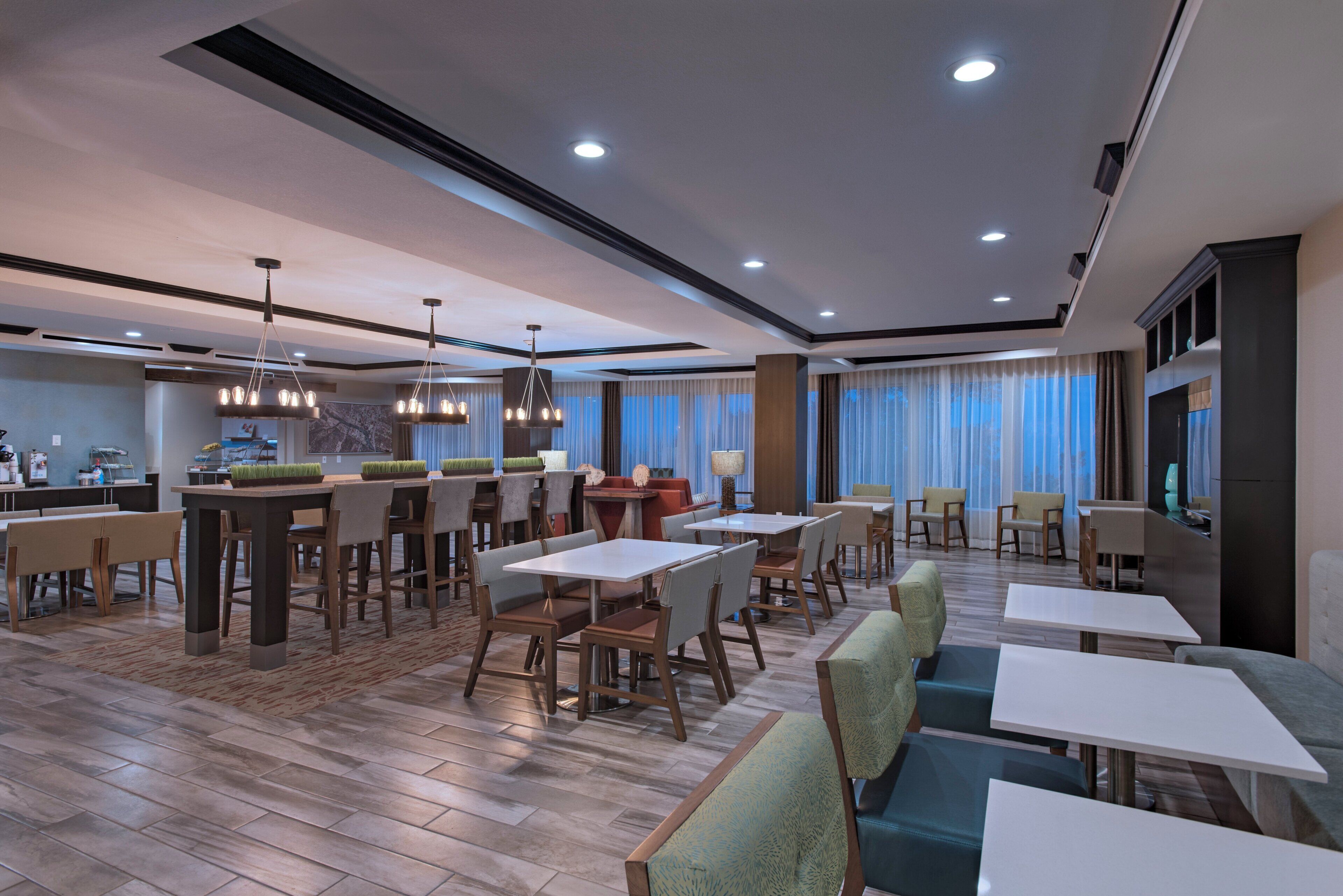 Une salle à manger d’hôtel avec plusieurs tables et chaises, un coin bar et une grande fenêtre avec des rideaux.