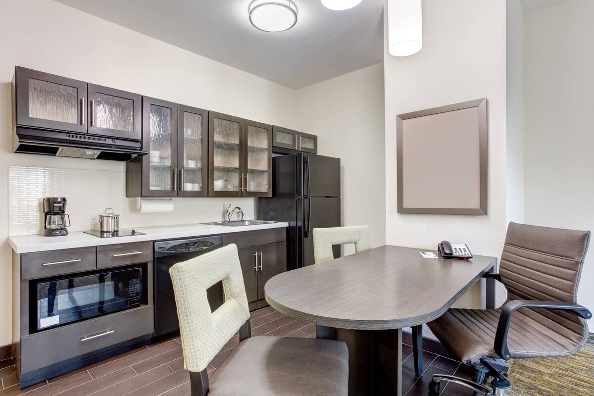 A modern kitchen with dark cabinets, a built-in oven, and a dining area with a large table and two chairs.