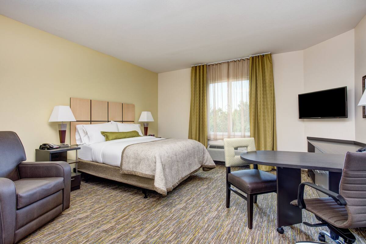 A hotel room with a bed, two chairs, a desk, a TV, and a window with curtains.
