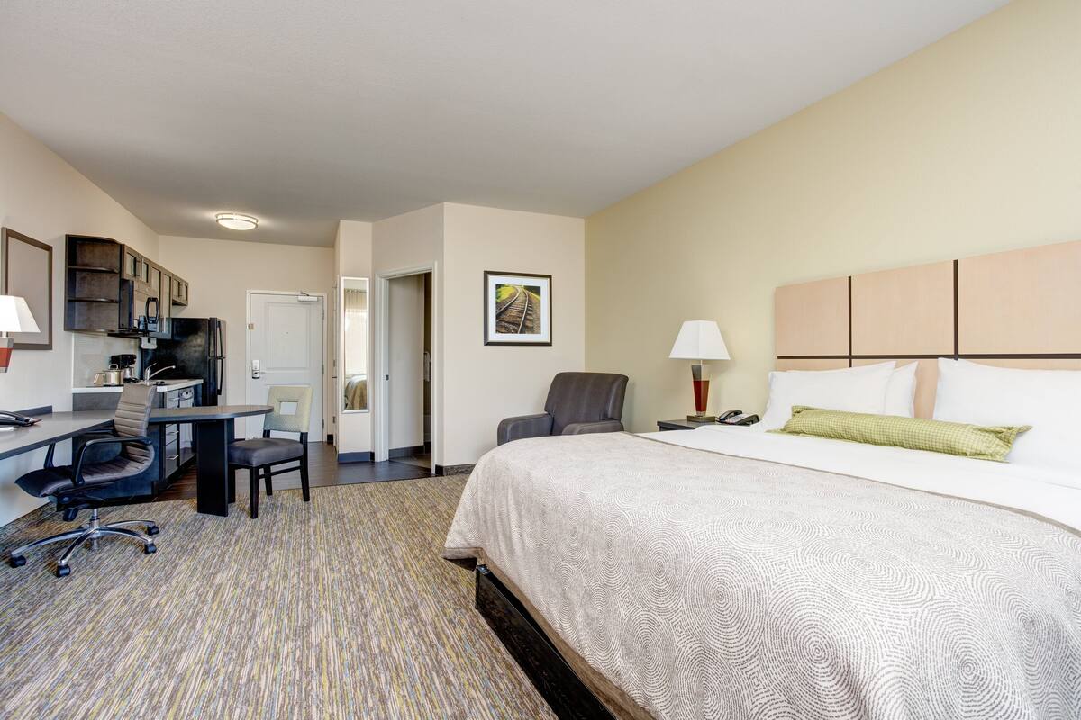 A hotel room with a bed, desk, and chair, a kitchenette, and a door leading to another room.
