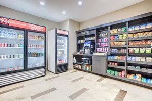 A self-service beverage and snack area with refrigerated drink dispensers, a microwave, and shelves stocked with various snacks and condiments. - Candlewood Suites Cut Off by IHG (Cut Off)