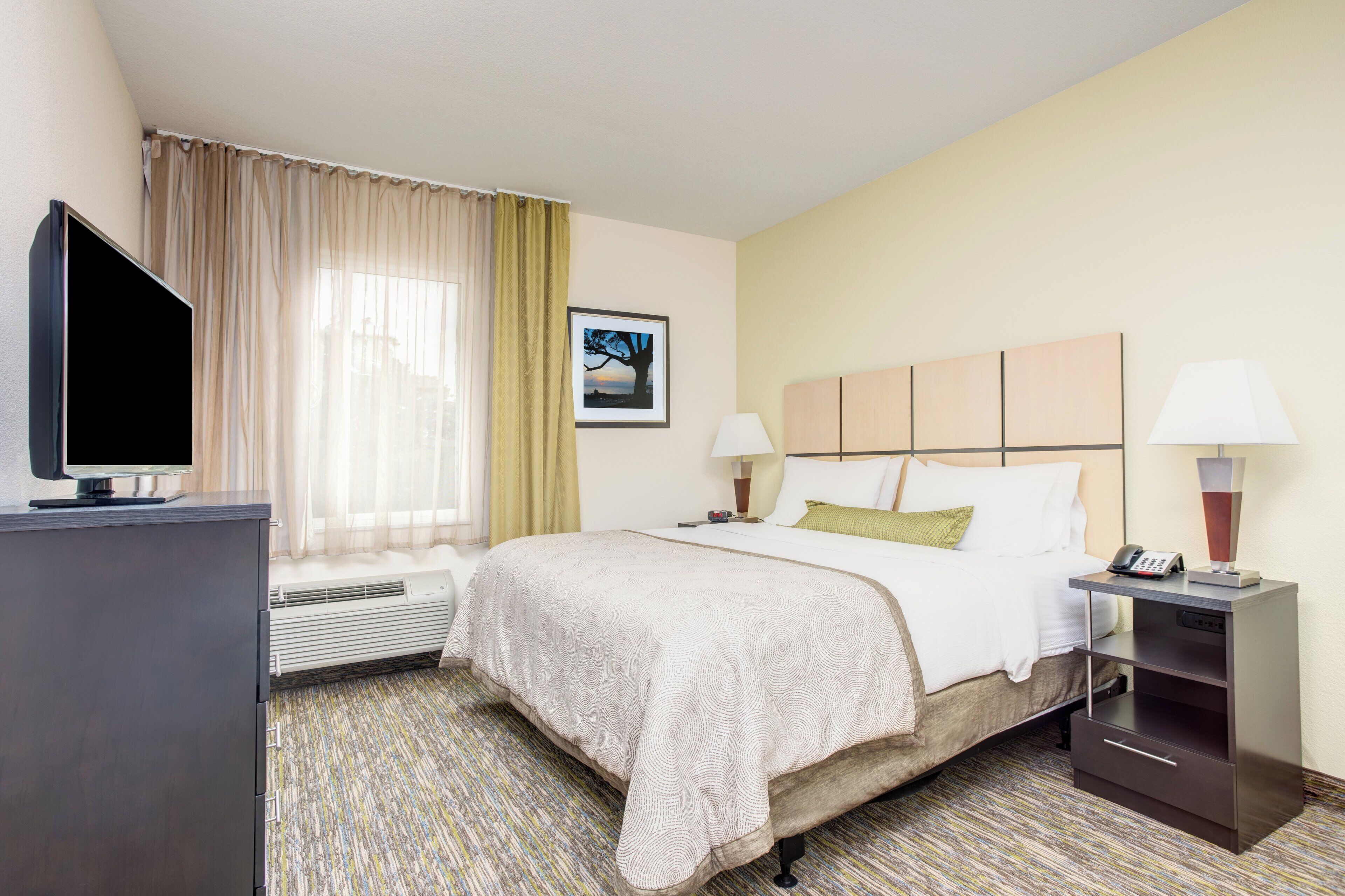 A hotel room with a bed, bedside lamps, a television, a picture on the wall, and a window with curtains.