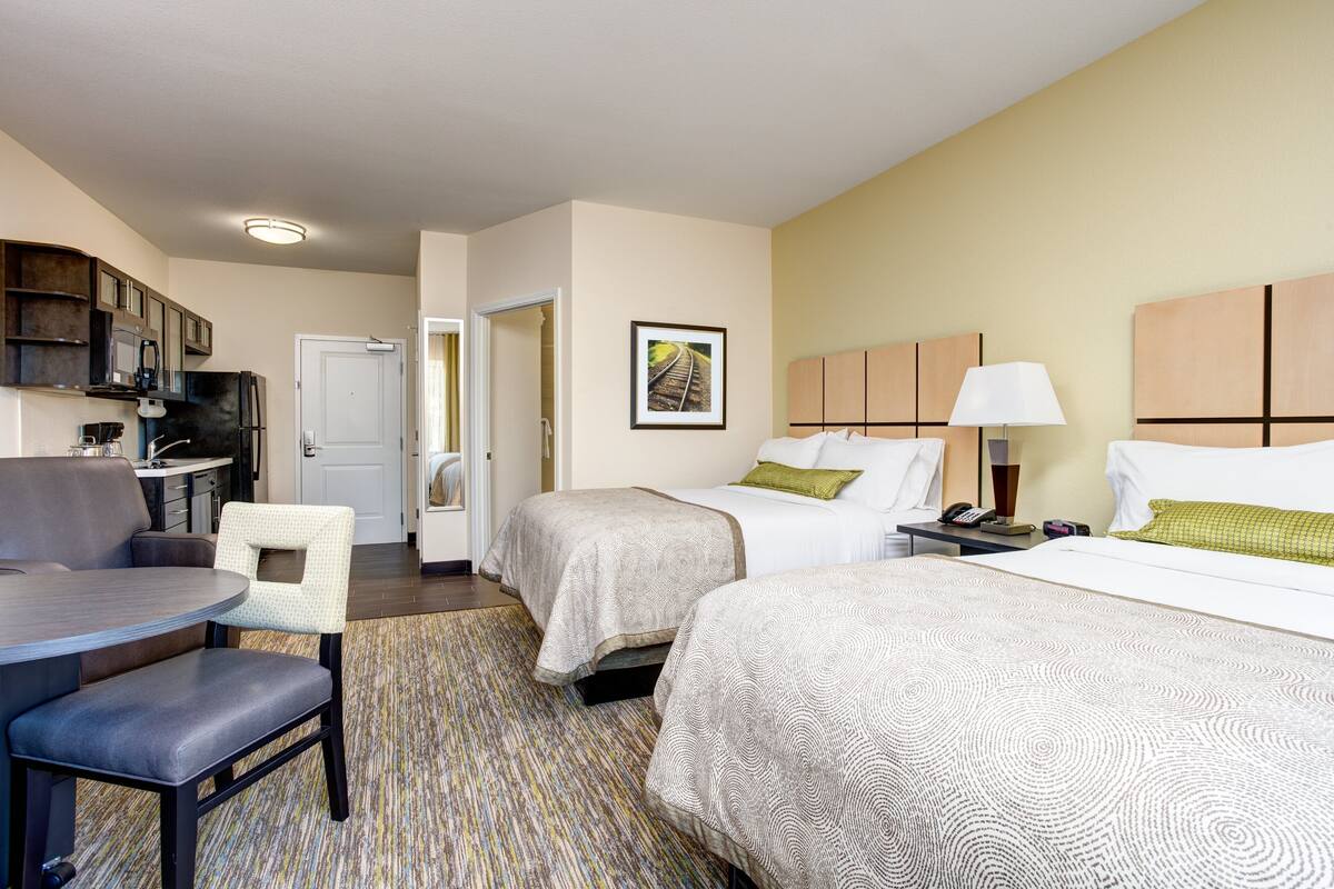 A hotel room with two beds, a desk, a chair, a kitchen area, and a door leading to another room.