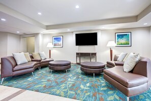 A hotel room with a sectional sofa, ottoman, flat-screen TV, and a small table with a lamp. - Candlewood Suites Cut Off by IHG (Cut Off)